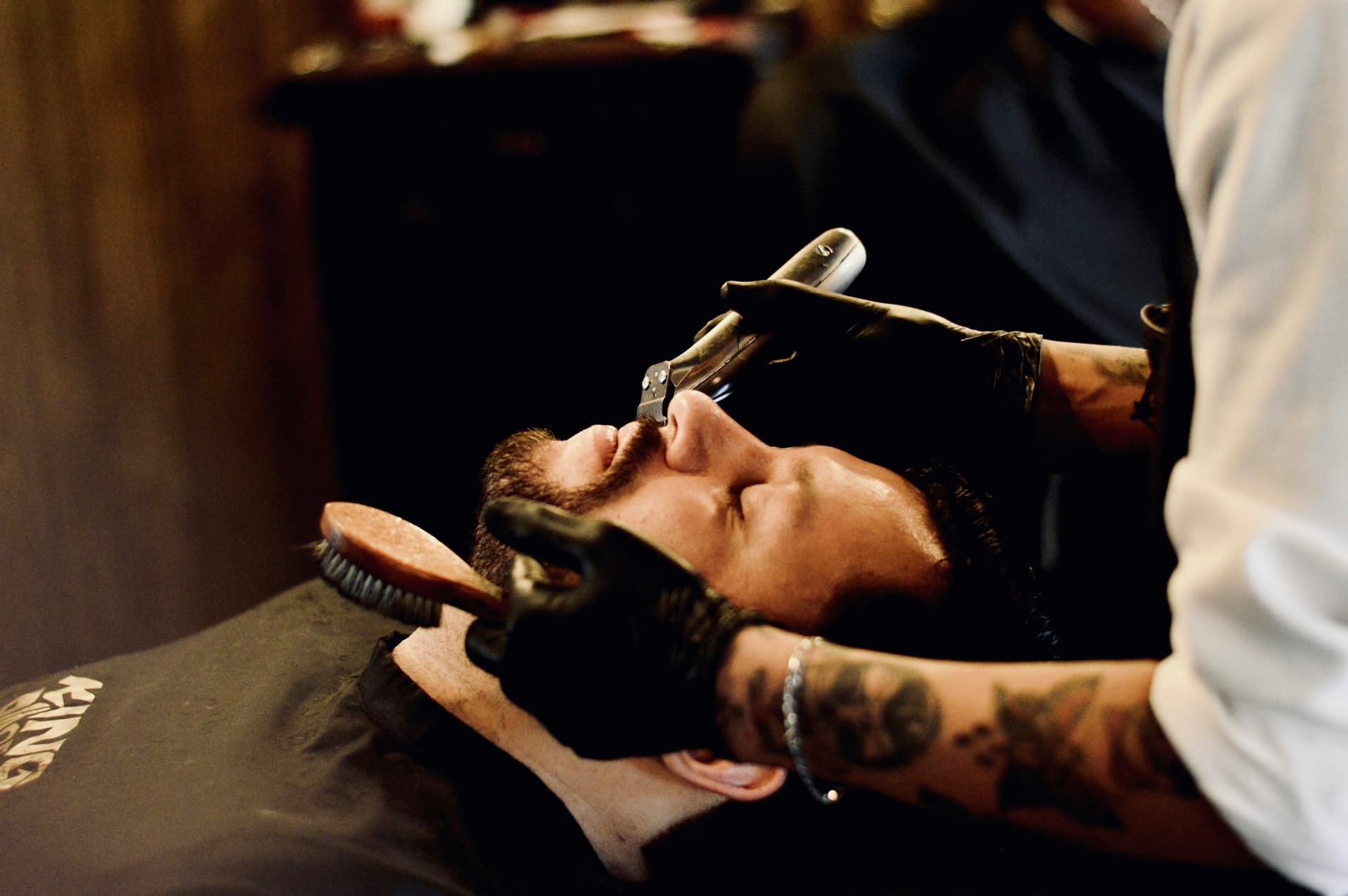 A barber holding a straight razor while grooming a reclined client in a dimly lit studio.